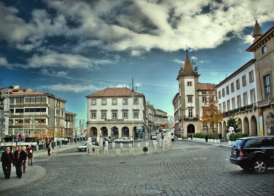 Praça na Covilhã. Está de dia, com o céu um pouco nublado, mas azul. Algumas pessoas andam no passeio. Alguns carros estacionados. A estrada é de pedra cinzenta, e os edifícios são brancos.